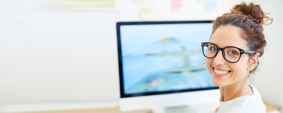 Frau mit Brille sitzt lächelnd vor Computerbildschirm im Büro