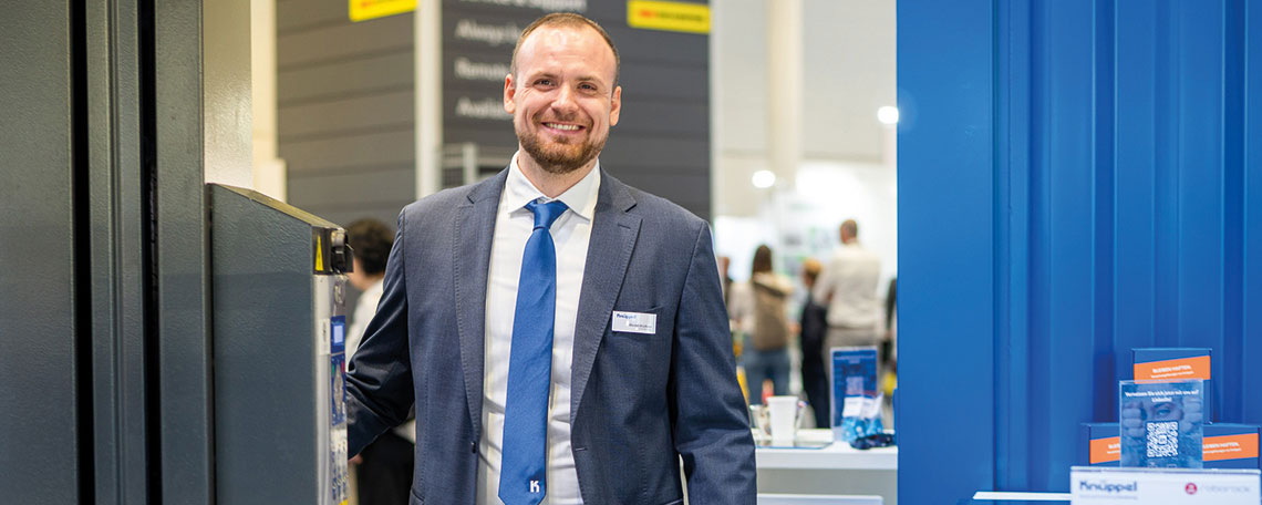 Ein Mann im Anzug steht an einem Messestand in einer Ausstellungshalle