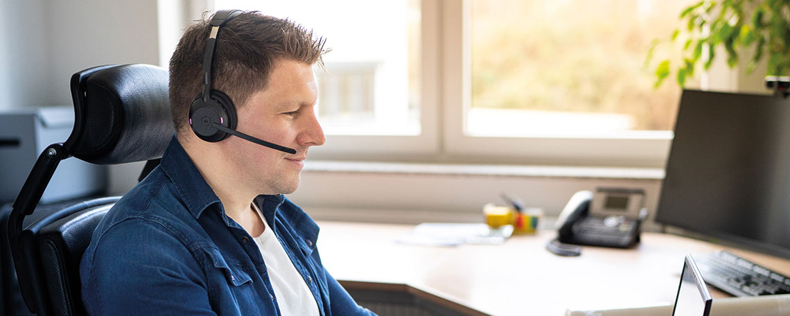 Ein Mann sitzt an einem Schreibtisch im Büro, trägt ein Headset und arbeitet am Computer